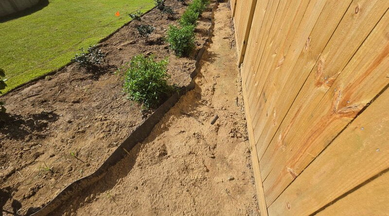Close-up of an HDPE root barrier trench running along a fence next to newly planted shrubs
