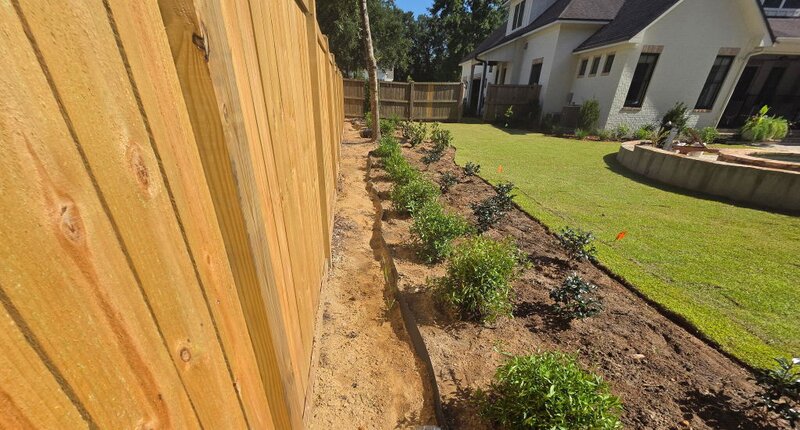 HDPE root barrier trench installed along a backyard fence line bordering a planted shrub bed
